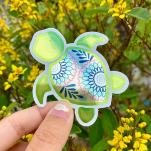 May include: A green and blue turtle sticker with a decorative pattern on its shell. The sticker has a shiny, iridescent finish.
