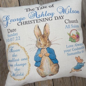 May include: A white pillow with a blue and gold design featuring a Peter Rabbit illustration. The text on the pillow reads "The Tale of George Ashley Wilson Christening Day Church All Saints Date Sunday 10.07.22 Even the smallest one can change the World Love Always your Godmother Samantha xx".