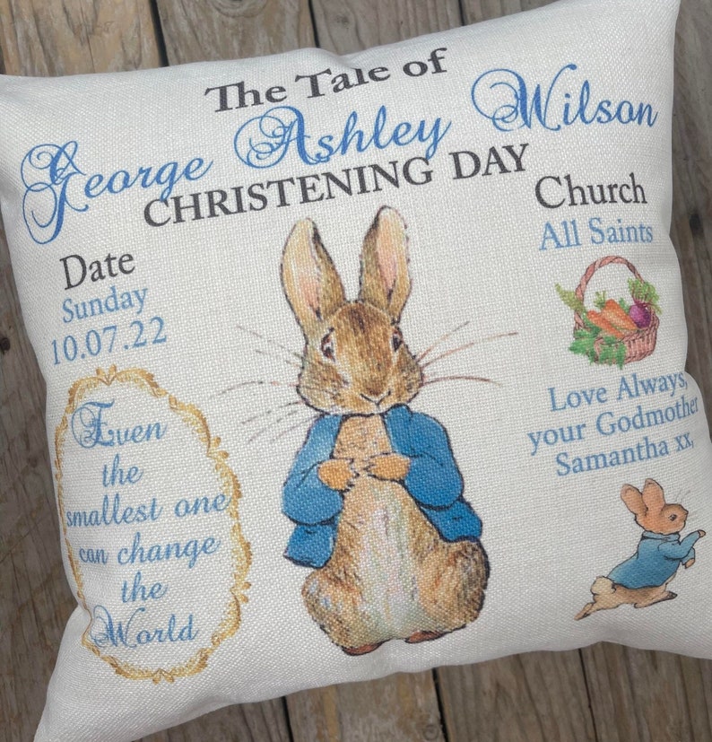 May include: A white pillow with a blue and gold design featuring a Peter Rabbit illustration. The text on the pillow reads "The Tale of George Ashley Wilson Christening Day Church All Saints Date Sunday 10.07.22 Even the smallest one can change the World Love Always your Godmother Samantha xx".