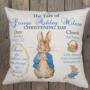 May include: A white pillow with a blue border featuring a Peter Rabbit illustration and the text "The Tale of George Ashley Wilson Christening Day", "Date Sunday 10.07.22 Church All Saints", and "Even the smallest one can change the World".  The text "Love Always, your Godmother Samantha xx" is also included.
