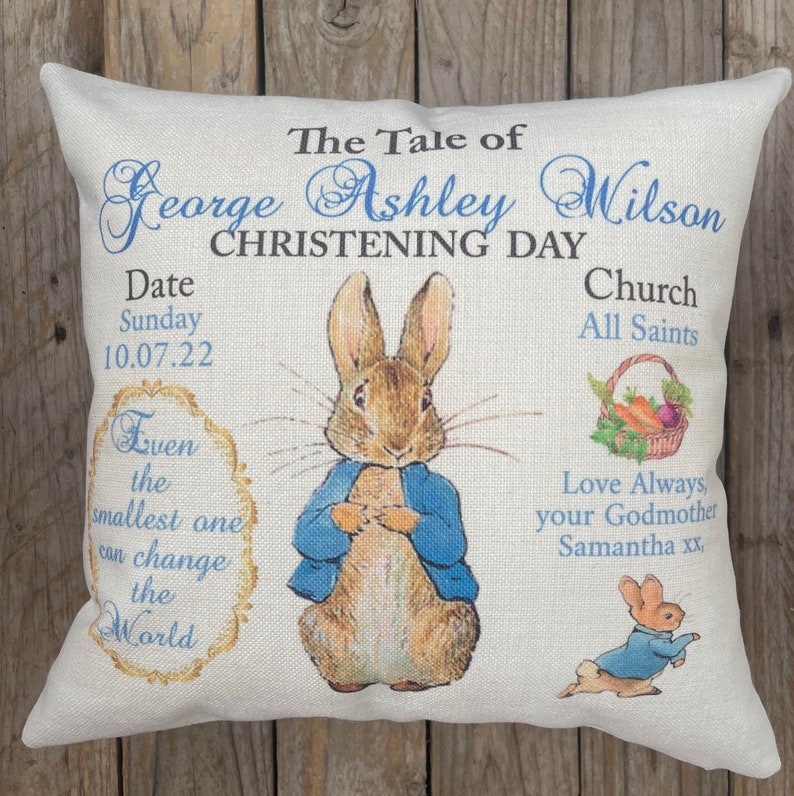 May include: A white pillow with a blue border featuring a Peter Rabbit illustration and the text "The Tale of George Ashley Wilson Christening Day", "Date Sunday 10.07.22 Church All Saints", and "Even the smallest one can change the World".  The text "Love Always, your Godmother Samantha xx" is also included.