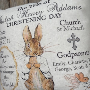 May include: A white pillow with a beige background featuring a brown rabbit illustration and the text "The Tale of Ralph Henry Addams Christening Day Church St Michael's Godparents Emily, Charlotte, T George, Scott & S".
