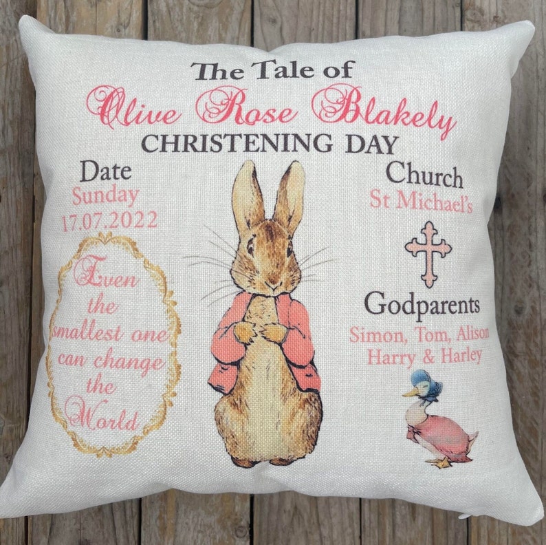 May include: A white pillow with a pink and brown illustration of Peter Rabbit. The pillow has the text "The Tale of Olive Rose Blakely Christening Day" and includes the date "Sunday 17.07.2022", the church "St Michael's", and the names of the godparents. The pillow also has the quote "Even the smallest one can change the world".