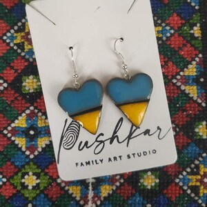 May include: A pair of heart-shaped earrings with blue, yellow, and black enamel. The earrings are on a white card with the text "Pushkar Family Art Studio".