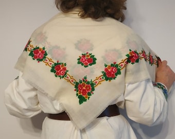 May include: A white embroidered scarf with red and green floral designs. The scarf is folded in half and draped over the shoulders of a person wearing a white shirt and blue jeans.