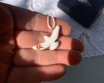 Könnte beinhalten: Ein weißer Keramik-Taubenanhänger mit roten und schwarzen Akzenten, an einer cremefarbenen Kordel. Die Taube wird in einer Hand gehalten, mit einer schwarzen Schachtel im Hintergrund. Der Anhänger ist ein Friedenssymbol.