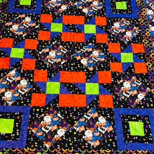 May include: A Halloween-themed quilt with a patchwork design featuring orange, blue, and green squares. The centre of the quilt is a black fabric with a repeating pattern of ghosts, pumpkins, and eyes. The border of the quilt is a black fabric with a repeating pattern of green and purple circles.