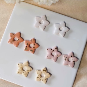 Peut inclure: Boucles d'oreilles en forme de fleurs dans des tons pêche, rose et crème, avec des accents de feuilles d'or. Elles sont disposées sur une surface carrelée blanche, mettant en valeur leur design délicat.