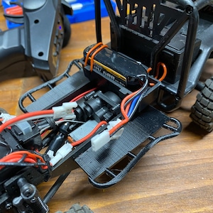 May include: A black and red remote control car with a roll cage and a battery pack. The car has a black chassis and red wires.