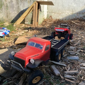 May include: A red toy truck with a flatbed trailer, hauling a smaller red toy truck. The larger truck has the words "Atlas 6x6" printed on the side. Both trucks are on a bed of wood chips.