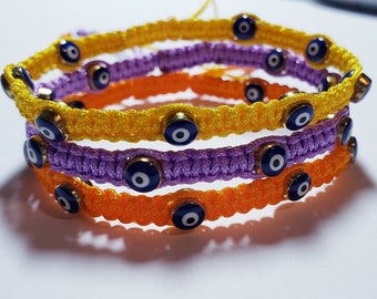 May include: Three colorful bracelets with blue evil eye charms. The bracelets are made of yellow, orange, and purple string.