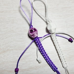 May include: Two skull-shaped keychains, one purple and one white, with purple and white cords. The cords are tied in a knot at the top and have small beads along the length.
