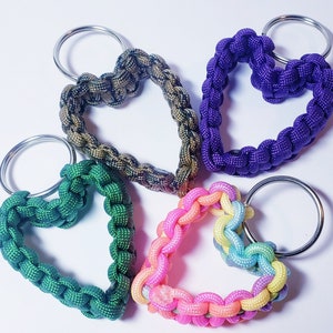 May include: Five heart-shaped keychains in various colors: green, purple, pink, and a camouflage pattern. Each keychain is made of woven cord and attached to a silver metal ring. The keychains are displayed on a white surface.