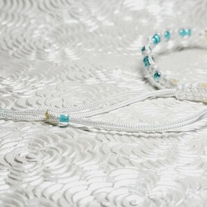 May include: White macrame bracelet with turquoise beads and a gold-colored four-leaf clover charm. The bracelet has adjustable white cords with turquoise accents. A delicate piece of jewelry.