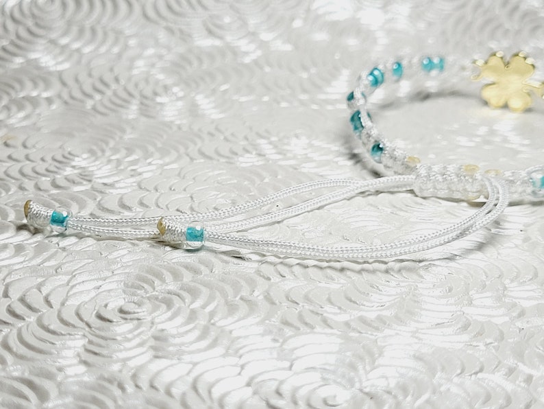 May include: White macrame bracelet with turquoise beads and a gold-colored four-leaf clover charm. The bracelet has adjustable white cords with turquoise accents. A delicate piece of jewelry.