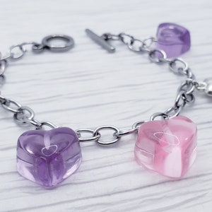 May include: A silver chain bracelet with a toggle clasp features five heart-shaped charms. Two of the charms are pink and three are purple.