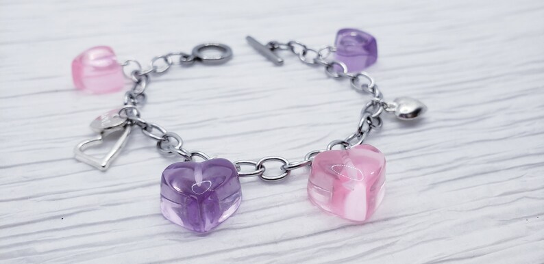 May include: A silver chain bracelet with a toggle clasp features five heart-shaped charms. Two of the charms are pink and three are purple.