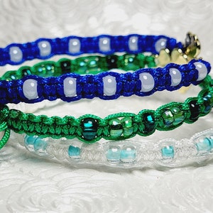 May include: Close-up of three macrame bracelets in blue, green, and white. The blue bracelet has white beads, the green has green and black beads, and the white has blue beads. Each bracelet has a gold-colored charm.
