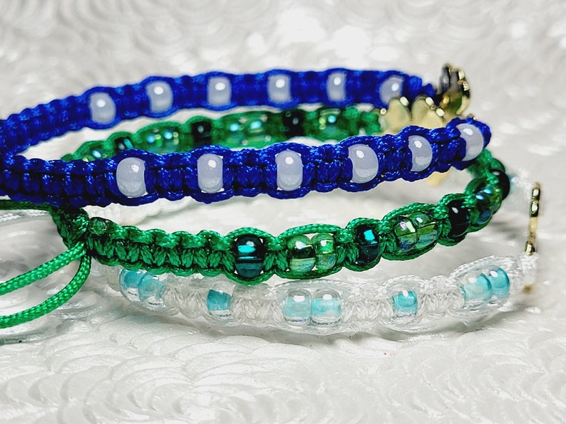 May include: Close-up of three macrame bracelets in blue, green, and white. The blue bracelet has white beads, the green has green and black beads, and the white has blue beads. Each bracelet has a gold-colored charm.