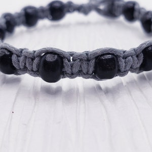 Puede incluir: Una pulsera de macram&eacute; gris con cuentas de madera negras. La pulsera est&aacute; hecha de cord&oacute;n gris, con las cuentas espaciadas uniformemente alrededor de la circunferencia. La pulsera se muestra sobre una superficie blanca.