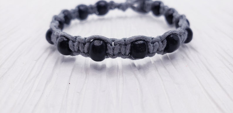 Puede incluir: Una pulsera de macram&eacute; gris con cuentas de madera negras. La pulsera est&aacute; hecha de cord&oacute;n gris, con las cuentas espaciadas uniformemente alrededor de la circunferencia. La pulsera se muestra sobre una superficie blanca.