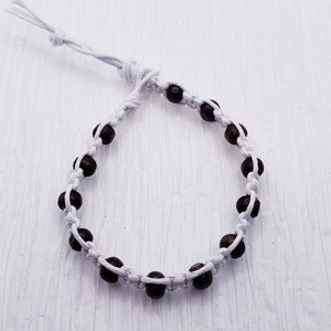 Puede incluir: Una pulsera hecha a mano con cuentas marrones oscuras ensartadas en un cord&oacute;n blanco. La pulsera es ajustable con un cierre de nudo corredizo. Las cuentas est&aacute;n espaciadas uniformemente, creando un dise&ntilde;o simple y elegante. La pulsera se muestra sobre una superficie blanca.