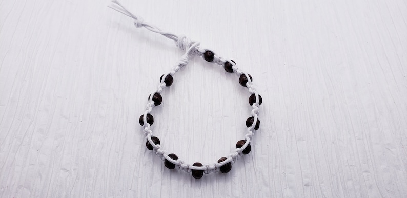 Puede incluir: Una pulsera hecha a mano con cuentas marrones oscuras ensartadas en un cord&oacute;n blanco. La pulsera es ajustable con un cierre de nudo corredizo. Las cuentas est&aacute;n espaciadas uniformemente, creando un dise&ntilde;o simple y elegante. La pulsera se muestra sobre una superficie blanca.
