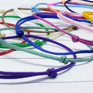 May include: A collection of colorful string bracelets with adjustable knots. The bracelets are made of thin, braided cord and come in a variety of colors, including purple, red, green, blue, yellow, pink, and white.