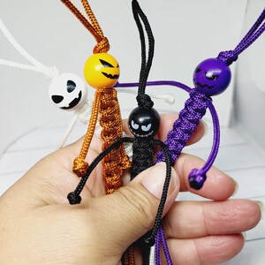 May include: Four Halloween-themed keychains with different coloured cords and wooden bead faces. The keychains feature a pumpkin, a ghost, a black cat, and a purple monster.