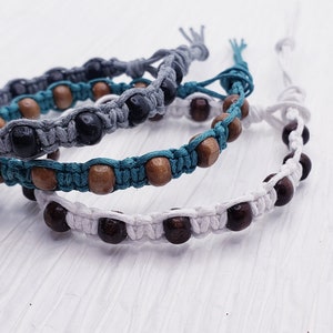 Puede incluir: Tres pulseras de macram&eacute; hechas a mano. Una es gris con cuentas negras, otra es turquesa con cuentas de madera y la tercera es blanca con cuentas marrones oscuras. Las pulseras se exhiben sobre una superficie blanca texturizada.