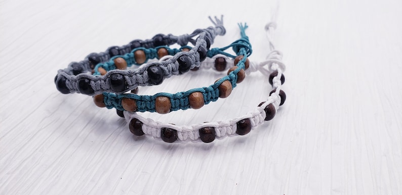 Puede incluir: Tres pulseras de macram&eacute; hechas a mano. Una es gris con cuentas negras, otra es turquesa con cuentas de madera y la tercera es blanca con cuentas marrones oscuras. Las pulseras se exhiben sobre una superficie blanca texturizada.