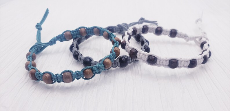 Puede incluir: Tres pulseras hechas a mano con cuentas de madera y cord&oacute;n anudado en verde azulado, gris y blanco. Las cuentas de madera son marrones y marr&oacute;n oscuro. Las pulseras son ajustables y tienen un dise&ntilde;o sencillo.