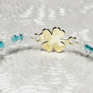 May include: A white macrame bracelet with turquoise beads and a gold-colored four-leaf clover clasp. The bracelet is made of woven white cord and features small, round turquoise beads interspersed along its length. The clasp is a detailed four-leaf clover.