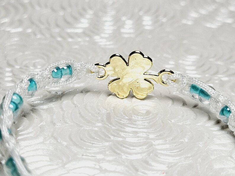 May include: A white macrame bracelet with turquoise beads and a gold-colored four-leaf clover clasp. The bracelet is made of woven white cord and features small, round turquoise beads interspersed along its length. The clasp is a detailed four-leaf clover.