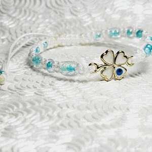 May include: White macrame bracelet with turquoise beads and a gold-colored four-leaf clover charm with a blue eye. The bracelet is adjustable and sits on a textured white surface.