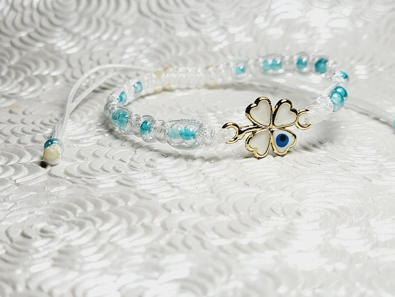 May include: White macrame bracelet with turquoise beads and a gold-colored four-leaf clover charm with a blue eye. The bracelet is adjustable and sits on a textured white surface.