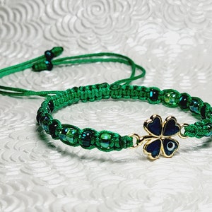 May include: A green macrame bracelet with iridescent beads and a gold-colored four-leaf clover charm. The clover has a dark blue enamel finish and a small evil eye detail. The bracelet has an adjustable sliding knot closure.