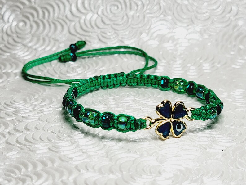 May include: A green macrame bracelet with iridescent beads and a gold-colored four-leaf clover charm. The clover has a dark blue enamel finish and a small evil eye detail. The bracelet has an adjustable sliding knot closure.