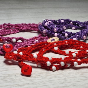 May include: Three crocheted bracelets with white beads and heart-shaped buttons. The bracelets are red, pink, and purple.