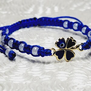 May include: A blue macrame bracelet with white beads and a gold-colored four-leaf clover charm. The clover has a blue and black enamel design with an evil eye in the center. The bracelet is adjustable.
