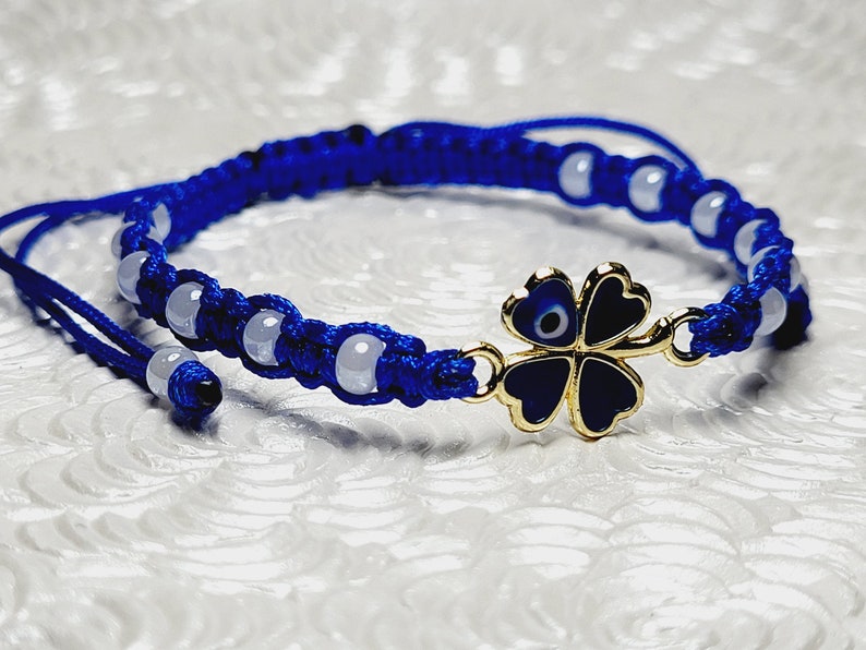 May include: A blue macrame bracelet with white beads and a gold-colored four-leaf clover charm. The clover has a blue and black enamel design with an evil eye in the center. The bracelet is adjustable.
