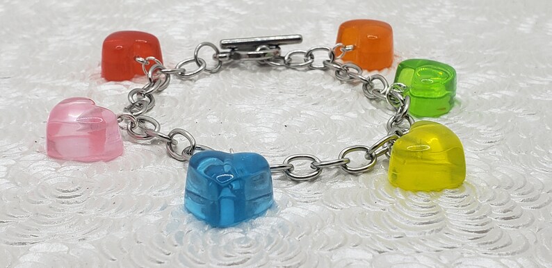 May include: A silver chain bracelet with five heart-shaped charms. The charms are made of clear plastic and are in the colors pink, orange, green, yellow, and blue.