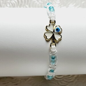 May include: White macrame bracelet with light blue beads and a gold-colored four-leaf clover charm with a blue eye in the center. The bracelet is displayed on a white surface.