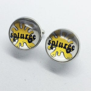 Può includere: Un paio di gemelli d'argento con un design a spruzzo giallo e nero. I gemelli presentano la parola "splurge" in lettere nere.