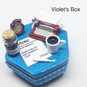 May include: A blue hexagonal box with a white ribbon and a white and red typewriter, a cup of coffee, a bottle of ink, a memo, and a clock. The memo reads "Memo to all employees Lunch breaks are now extended to 1 hour. FRHart".