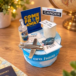 Puede incluir: Un expositor circular con un cartel del musical Come From Away, un cartel de bienvenida a Gander, una botella miniatura de Screech, un avión de plata, un pez azul pequeño y una tarjeta For the Lions Club. Base azul con el texto You are here.