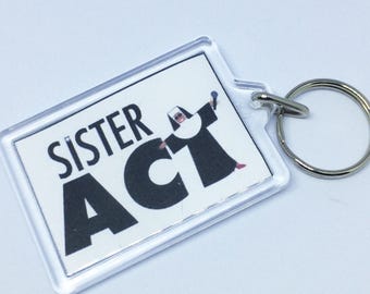 May include: A clear plastic keychain with a white background and black text that reads "Sister Act". A black and white image of a nun holding a microphone is in the background.