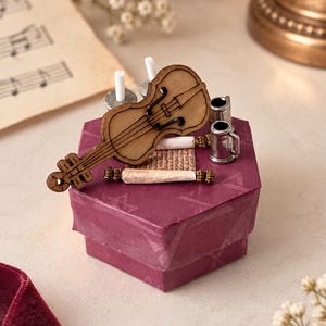 Peut inclure: Une boîte cadeau hexagonale violette surmontée d'un violon miniature en bois, d'une petite tasse en métal argenté et d'un minuscule parchemin roulé avec du texte visible. La boîte est entourée d'un fin ruban violet translucide.