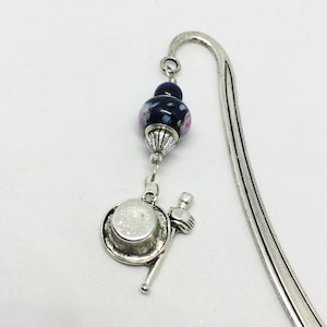 May include: A silver-toned metal bookmark featuring a curved hook top. A dark blue glass bead with floral patterns and a silver-toned metal cap hangs from the hook, attached to a charm shaped like a top hat and a walking cane in silver-toned metal.