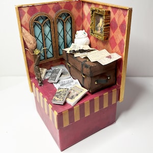 Może przedstawiać: Miniaturowa diorama pokoju z kufrem, książkami i miotłą. Ściany są czerwone z rombowym wzorem. Biała sowa siedzi na kufrze. Widoczne są dwa łukowate okna i oprawiony obraz. Podstawa jest czerwona ze złotymi paskami.